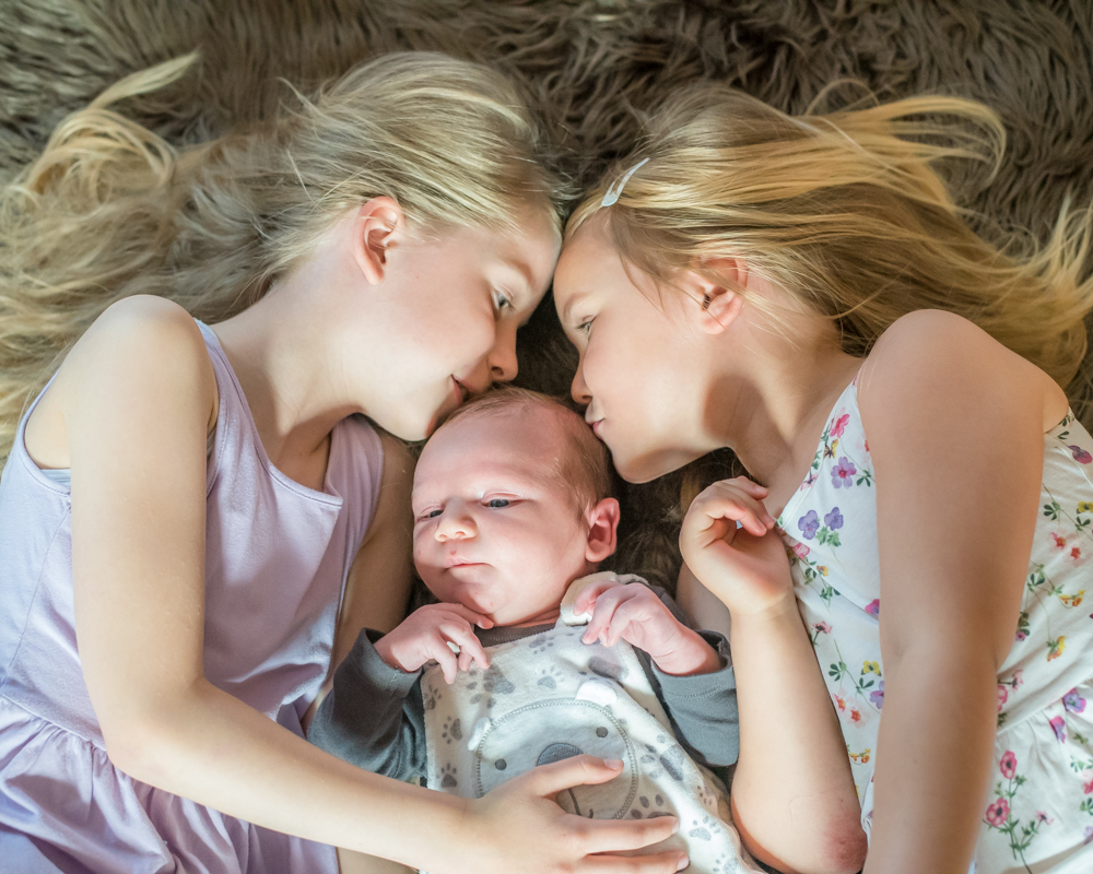 Sisters kissing baby Adam, newborn photographer Cumbria